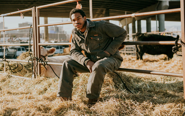 Man sits against fence wearing coveralls and posing in a cattle tie out with hay.